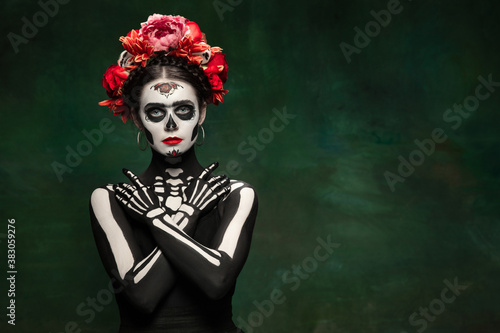 Bright. Young girl like Santa Muerte Saint death or Sugar skull with bright make-up. Portrait isolated on dark green studio background with copyspace. Celebrating Halloween or Day of the dead.