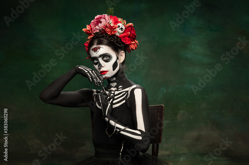 Snake queen. Young girl like Santa Muerte Saint death or Sugar skull with bright make-up. Portrait isolated on dark green studio background with copyspace. Celebrating Halloween or Day of the dead.