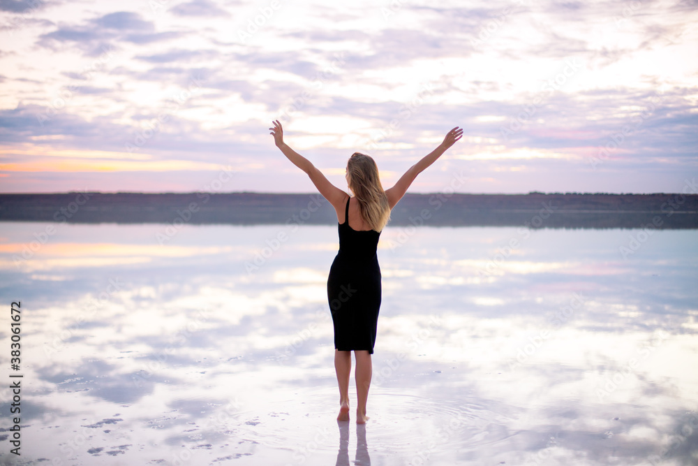 beautiful girl with brown hair and a slim figure in a black dress is staying with raised hands on a salty pink color lake at a vanilla sunset light. view from the back