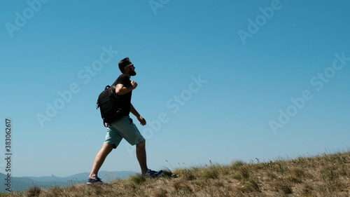 Young bearded guy in black T-shirt, blue shorts and with backpack on his back climbs up the hill. Man conquers high mountains. Grass grows on the mountains and man walks forward, side view.