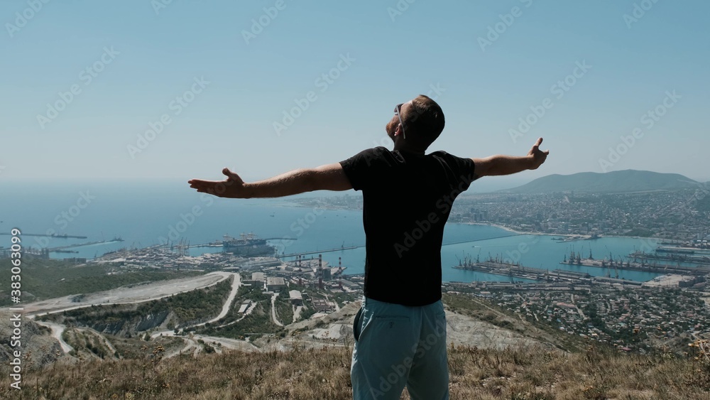 Man on the top om hill and spread his arms in different directions. Young bearded man in black t-shirt and sunglasses enjoy the moment.