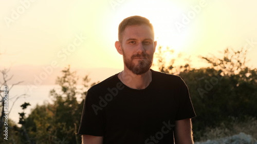 Portrait of handsome pretty bearded red-haired guy outside at sunset looking at camera. A man stands in black T-shirt with orange sky in the back. © Vit