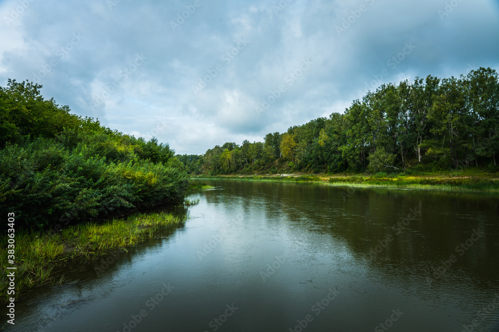 Calm river with trees on the shores in rainy summer day. Summer landscape.
