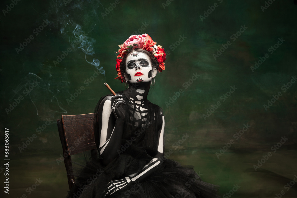 Smoking. Young girl like Santa Muerte Saint death or Sugar skull with bright make-up. Portrait isolated on dark green studio background with copyspace. Celebrating Halloween or Day of the dead. Smoking. Young girl like Santa Muerte Saint death or Sugar skull with bright make-up. Portrait isolated on dark green studio background with copyspace. Celebrating Halloween or Day of the dead.