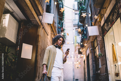 Positive curly male millennial standing on city street with mobile phone browsing web page with locations and destinations, smiling hipster guy holding smartphone reading messages and notifications © BullRun