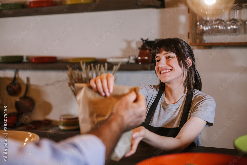 Waitress wearing apron serving customer at counter in restaurant. Young caucasian woman owner offering recycled paper bag with take away food to online client. Small business and service concept. Waitress wearing apron serving customer at counter in restaurant. Young caucasian woman owner offering recycled paper bag with take away food to online client. Small business and service concept.