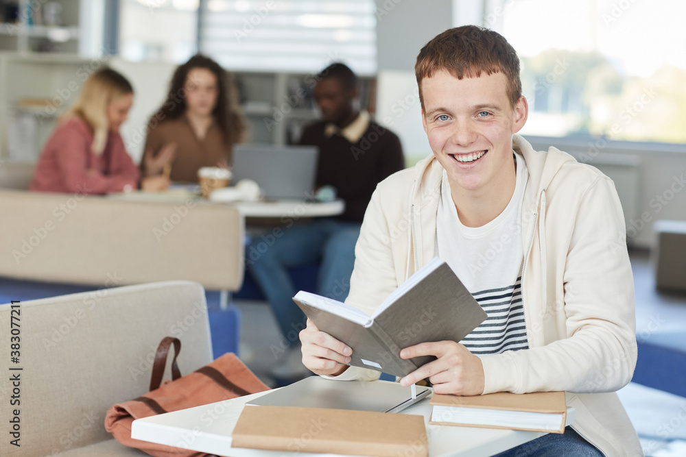 Portrait of smiling red haired boy looking at camera while studying in college library and holding textbook, copy space Portrait of smiling red haired boy looking at camera while studying in college library and holding textbook, copy space