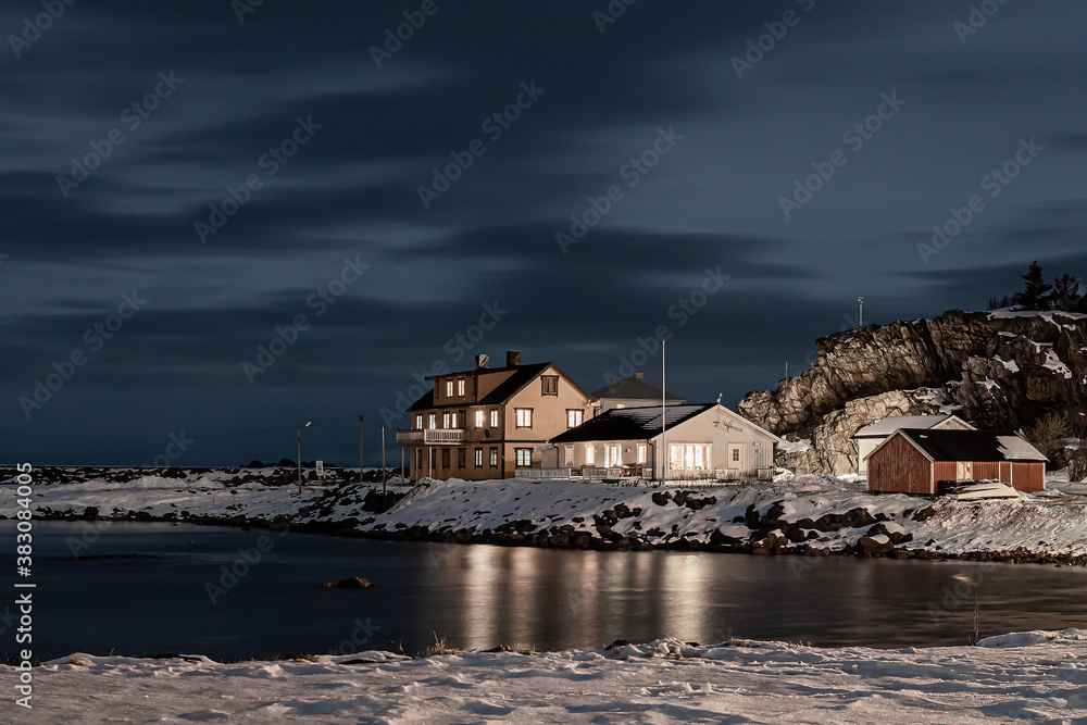 Wooden cottage on the shore of the sea at night in winter. View of harbour with wooden houses in winter season surrounded by snowy mountains