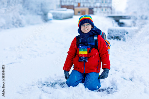 Gorgeous little school kid boy with glasses of elementary class walking to school during strong snowfall. Snowy streets in city. Child with backpack or satchel in colorful winter clothes