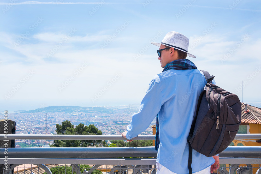 A young guy in a scarf, in hat and sunglasses with a backpack stands near the Temple of the Sacred Heart on Mount Tibidabo in Barcelona, Spain