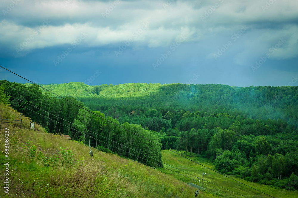 Dramatic landscape with thunder sky and green forest. Empty chairlift in ski resort in summer with green grass and no snow.