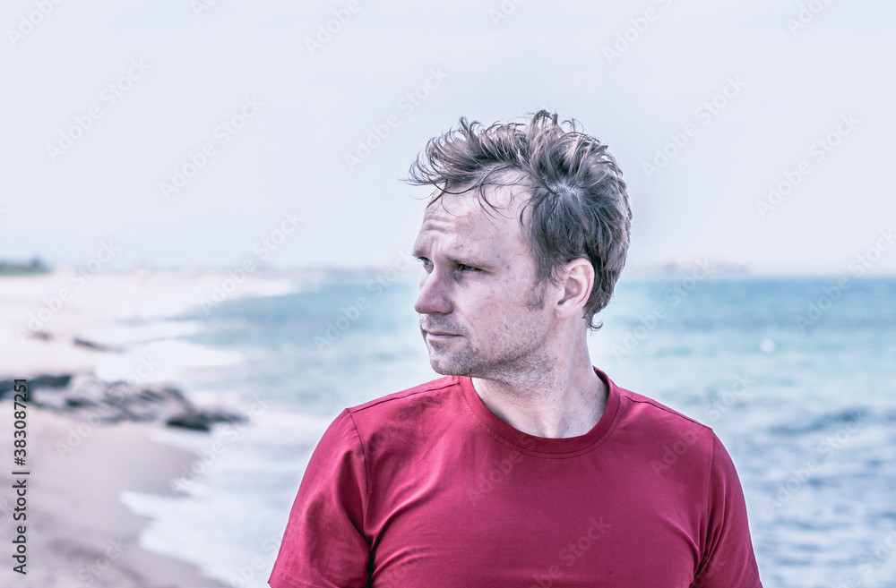 Fototapeta premium Serious handsome fair hair fluttering wind man red shirt, stubble face cheeks beard, wrinkled forehead sad, real skin, look far away thinking, blur sea skyline background BANNER. Life problems concept