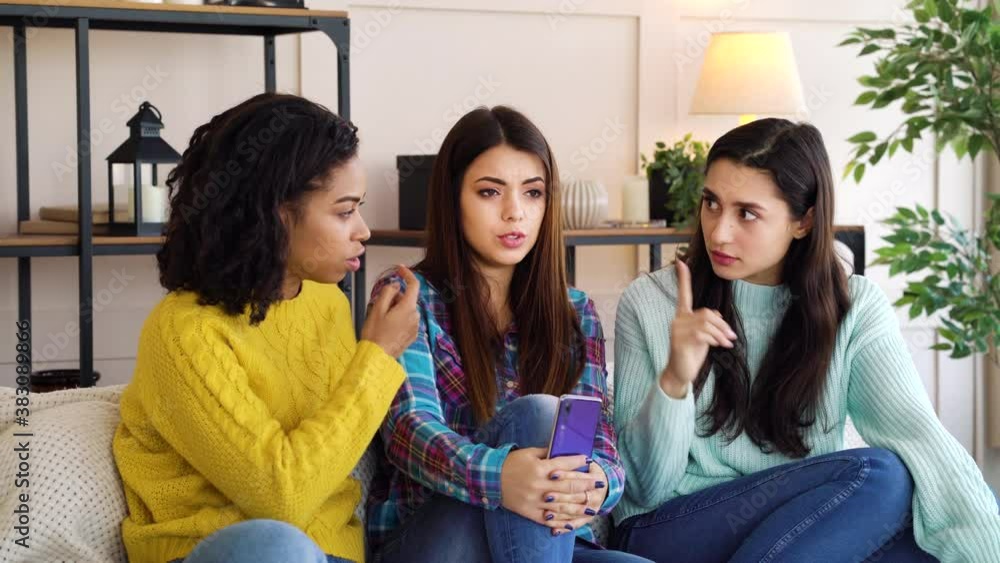 Young women in casual wear sitting in living room together, talking and gesturing, one of them holding smartphone. Multiracial friends having conversation. Concept of communication Young women in casual wear sitting in living room together, talking and gesturing, one of them holding smartphone. Multiracial friends having conversation. Concept of communication