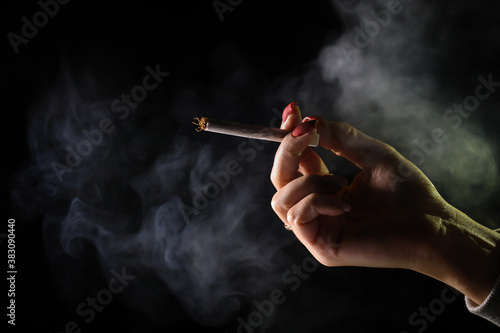 Woman hand holding cigarette in a smoke against black background. Smoking cannabis joint. Medical use.
