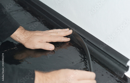A man is installing a black PVC skirting board along a white wallpapered wall and hardwood painted floor to conceal electric cables behind a plastic moulding.