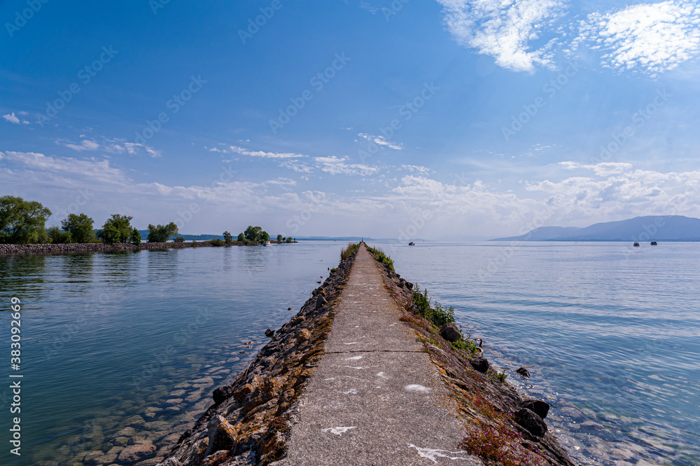  View on a very long dike leading to the Tene lighthouse on Lake Neuchatel in Switzerland. The blue sky is reflected in the calm water of the lake.