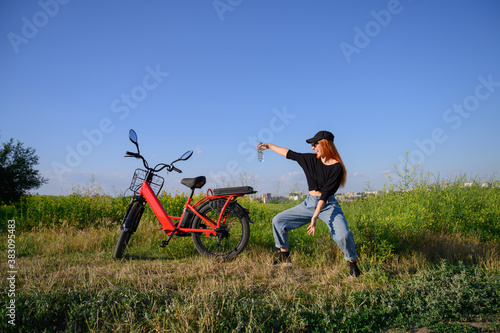 Beautiful caucasian girl stand near red electro bicycle at background of blue sky resting and enjoy after bike ride at sunny summer day. fitness, sport, people and healthy lifestyle concept