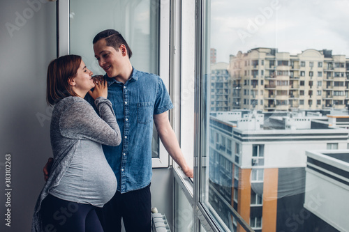 a man and a pregnant woman stand at home near an open window and talk. The husband hugs his wife, they stay at home and look at the street from the window. 