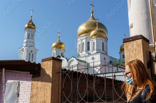 Caucasian woman in protective mask stands near fence with Christian European Church in the background. Coronavirus, health protection, quarantine, safety and pandemic concept