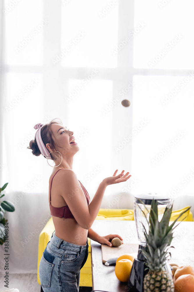 Selective focus of young housewife throwing kiwi near cutting board and blender Selective focus of young housewife throwing kiwi near cutting board and blender