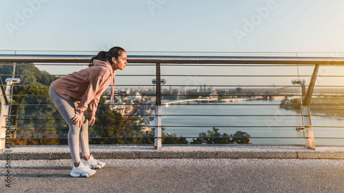 Young lady runner is resting after jogging on the bridge against city landscape