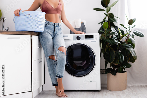 Cropped view of barefoot woman holding laundry basket near washing machine during housework at home