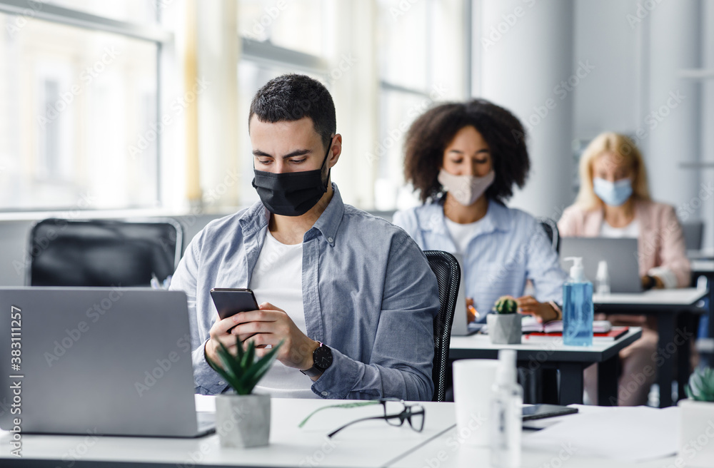 Modern gadgets for working with clients remotely from office after quarantine. Arabic young guy in protective mask typing on smartphone at table with laptop Modern gadgets for working with clients remotely from office after quarantine. Arabic young guy in protective mask typing on smartphone at table with laptop