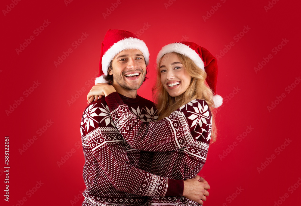 Expressing affection. Lovely young couple in Santa hats and Christmas sweaters hugging over red background Expressing affection. Lovely young couple in Santa hats and Christmas sweaters hugging over red background