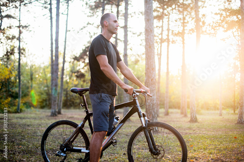 A caucasian man in a black t-shirt and shorts is staying with a black bike in a forest in the orange sunset light