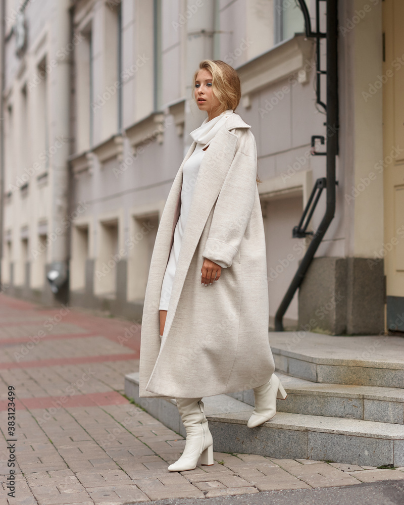 Elegant woman in white dress hessian boots and coat walking at city street. Fall autumn fashion