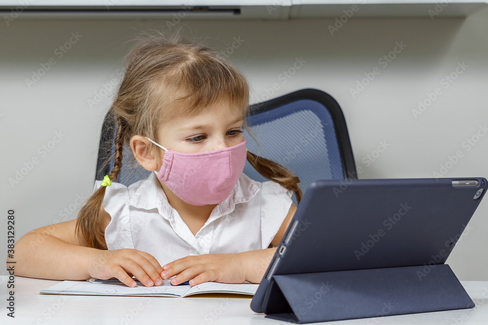 cute little girl in a pink protective mask sits at the table and looks into the tablet cute little girl in a pink protective mask sits at the table and looks into the tablet