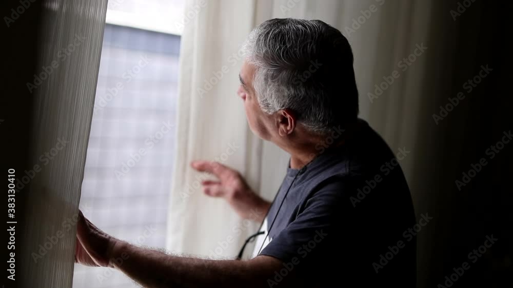 Elder man standing by window looking out surveilling noise Elder man standing by window looking out surveilling noise