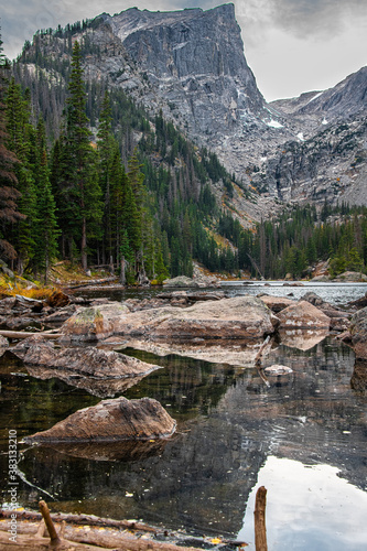 Dream Lake in Rocky Mountain National Park right outside of Estes Park. A mountain range and peaks are seen with a dark sky and rocks in a lake with reflections and trees. 