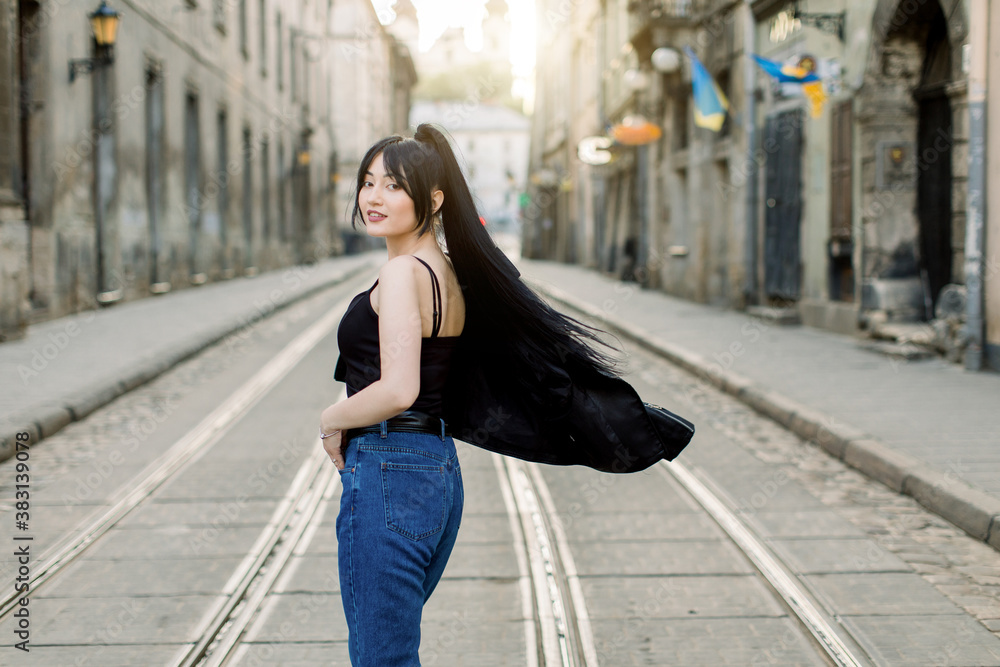Fototapeta premium Young beautiful Asian girl with ponytail black hair, wearing casual jeans and black top, turning to camera and posing while walking in the city on a sunny day. Outdoor fashion portrait