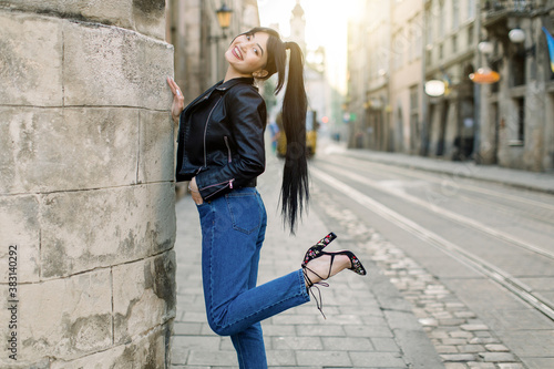 Beautiful young Asian model woman in fashionable casual black leather jacket and denim, and heels, standing near old building in ancient city center. Fashion model, urban lifestyle © sofiko14