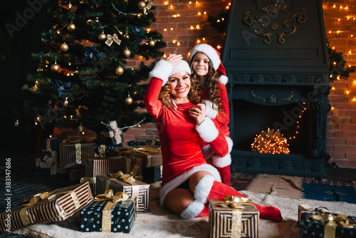 Beautiful young mother and daughter sitting on the floor next to a Christmas tree, hugging.