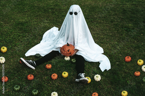 Lonely ghost sitting on a park surrounded by small pumpkins wearing glasses for halloween