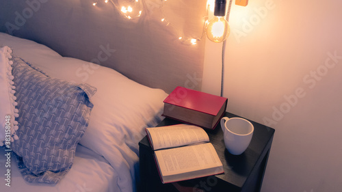 Leer un buen libro antes de dormir. Plano levemente elevado de mesa de noche junto a una cama con libros antiguos y una taza de café en un cálida confortable e iluminada habitación. 