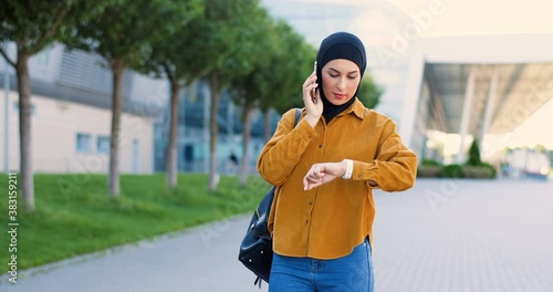 Muslim beautiful young woman in black hijab walking at street, talking on cellpphone and looking at hand watch to know time. Arabian pretty female waiting for meeting and speaking phone outdoors. photo