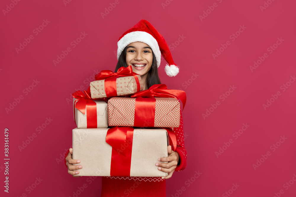 Funny indian latin preteen girl wears santa hat looking at camera holding many gifts boxes standing isolated on red background. Merry Christmas presents shopping sale, Happy 2021 New Year celebration. Funny indian latin preteen girl wears santa hat looking at camera holding many gifts boxes standing isolated on red background. Merry Christmas presents shopping sale, Happy 2021 New Year celebration.