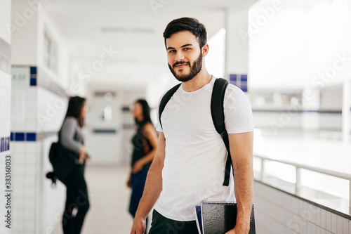 Portrait of young Brazilian student with backpack carrying books in college. Other students in the background.