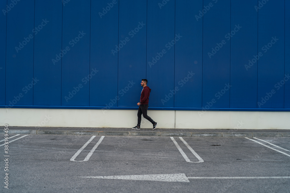 Chico joven con sudadera andando por delante de edificio azul Chico joven con sudadera andando por delante de edificio azul