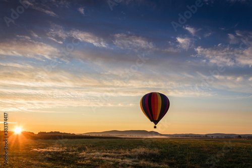 Hot Air Balloon in colorful rainbow stripes begins ascent over farm field as sun rises blue cloudy sky