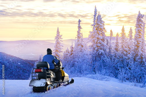 Man standing on snowy mountain near snowmobile enjoying view winter forest at sunset. Concept winter travel, sports, activity.