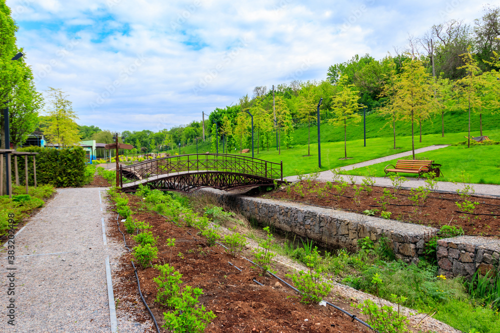 Beautiful view of the city park with arched footbridge across small river and young trees Beautiful view of the city park with arched footbridge across small river and young trees