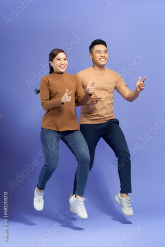 Energetic beautiful couple posing together on camera while running or jumping and pointing fingers on copyspace along background