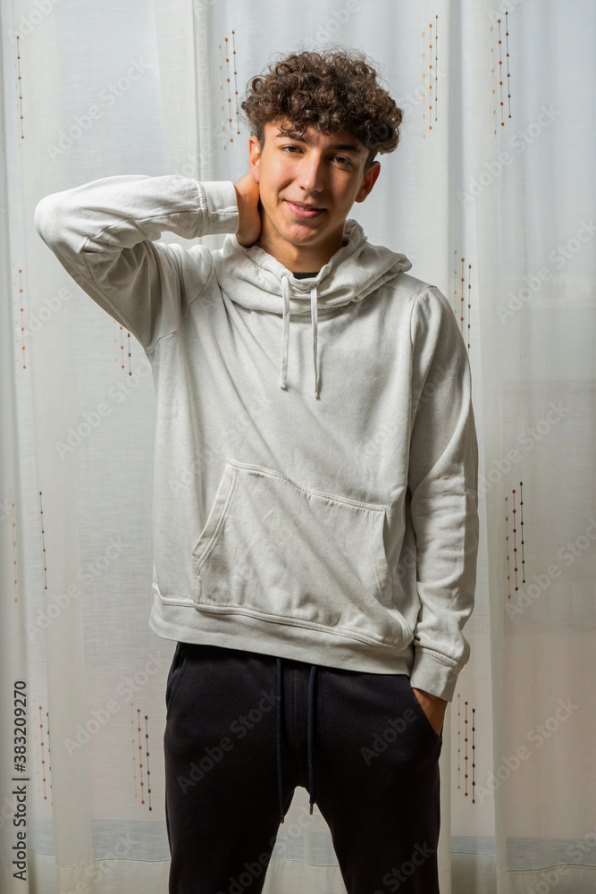 Attractive young man with curly hair wearing white sweatshirt posing on white curtains background Attractive young man with curly hair wearing white sweatshirt posing on white curtains background