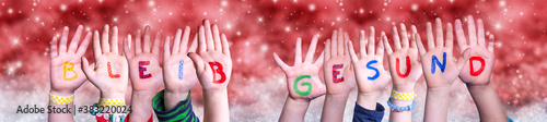 Children Hands Building Colorful German Word Bleib Gesund Means Stay Healthy. Red Snowy Christmas Winter Background With Snowflakes And Sparkling Lights