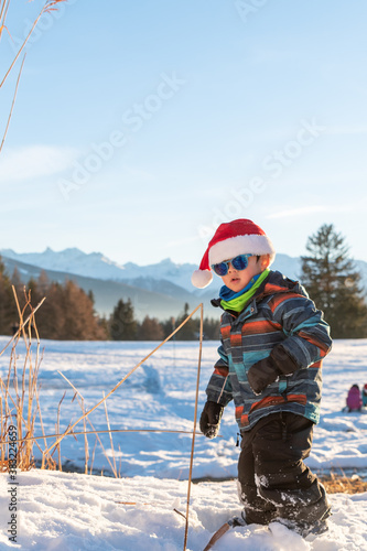 Boy with Santa red hat against a winter landscape background. Happy smiling child in the snow at Christmas standing beside tall plants outdoors. Crans Montana in Switzerland.