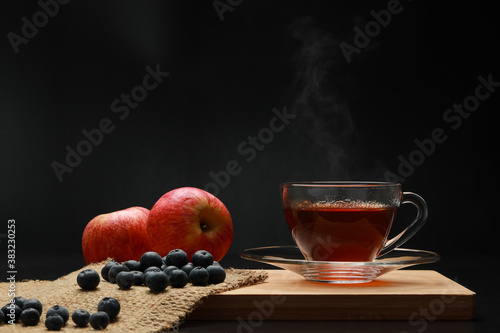 The hot tea fruits in glass cup with smoke and blueberry  apple on the wooden board as black background. Healthy drink concept. Dark tone.